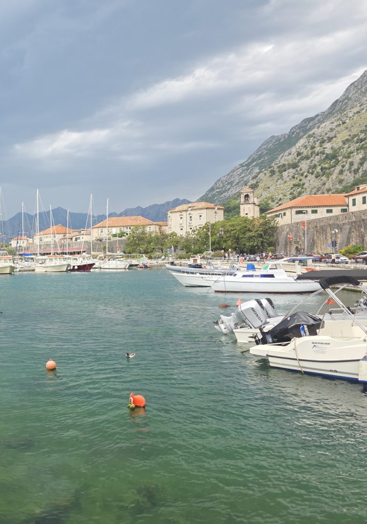 Small boats are docked in a turquoise harbor beside a stone old town and church tower. Steep mountains rise behind the waterfront under a cloudy sky.