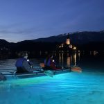 Clear kayak on Lake Bled at dusk with two people paddling over glowing blue water. Bled Island and the lit church reflect across the lake with dark mountains in the background.
