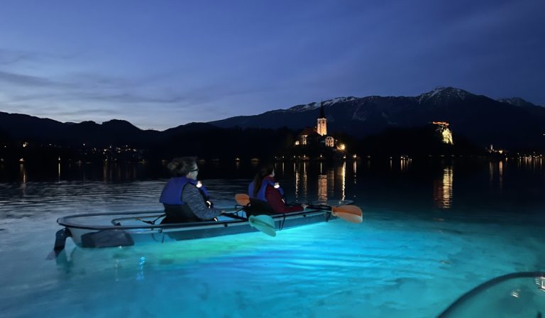 Clear kayak on Lake Bled at dusk with two people paddling over glowing blue water. Bled Island and the lit church reflect across the lake with dark mountains in the background.