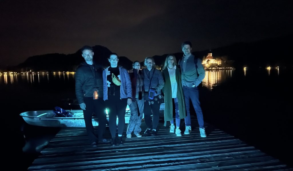 Six people stand on a wooden dock at night after kayaking on Lake Bled. A clear kayak glows blue beside them and Bled Island shines across the dark water in the background.