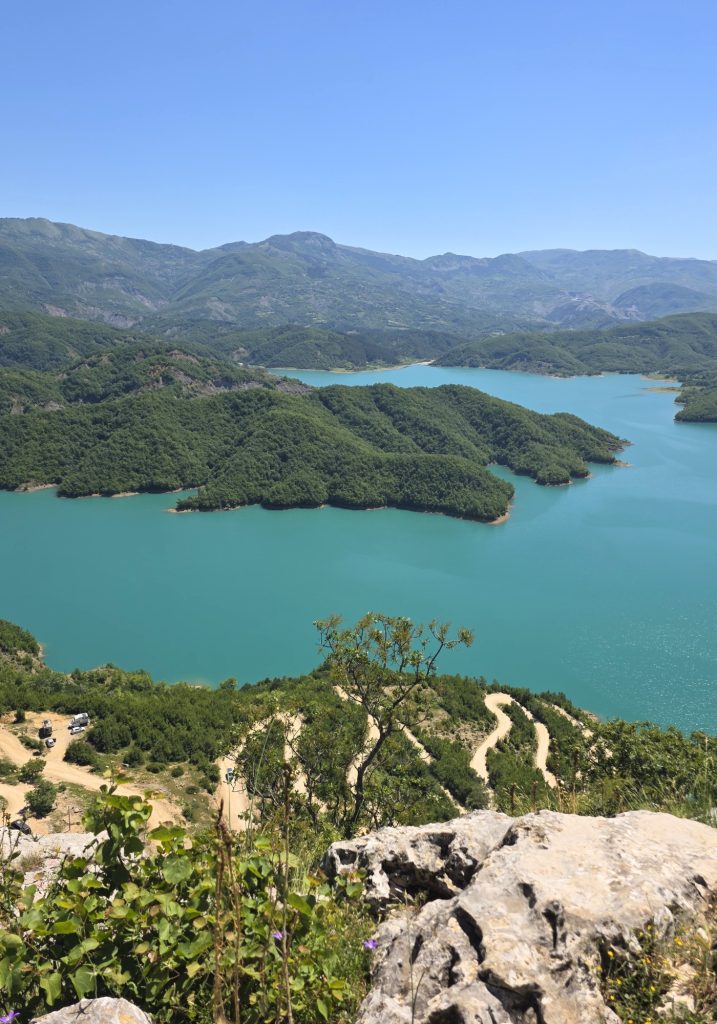 Turquoise lake winds between forested hills and mountains under a clear blue sky. A rocky overlook and dirt road in the foreground show the steep viewpoint above the water.