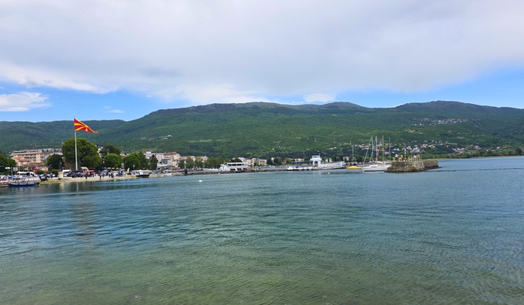 Lake Ohrid stretches across the foreground with boats at the marina a North Macedonian flag on shore and green mountains beyond the town.