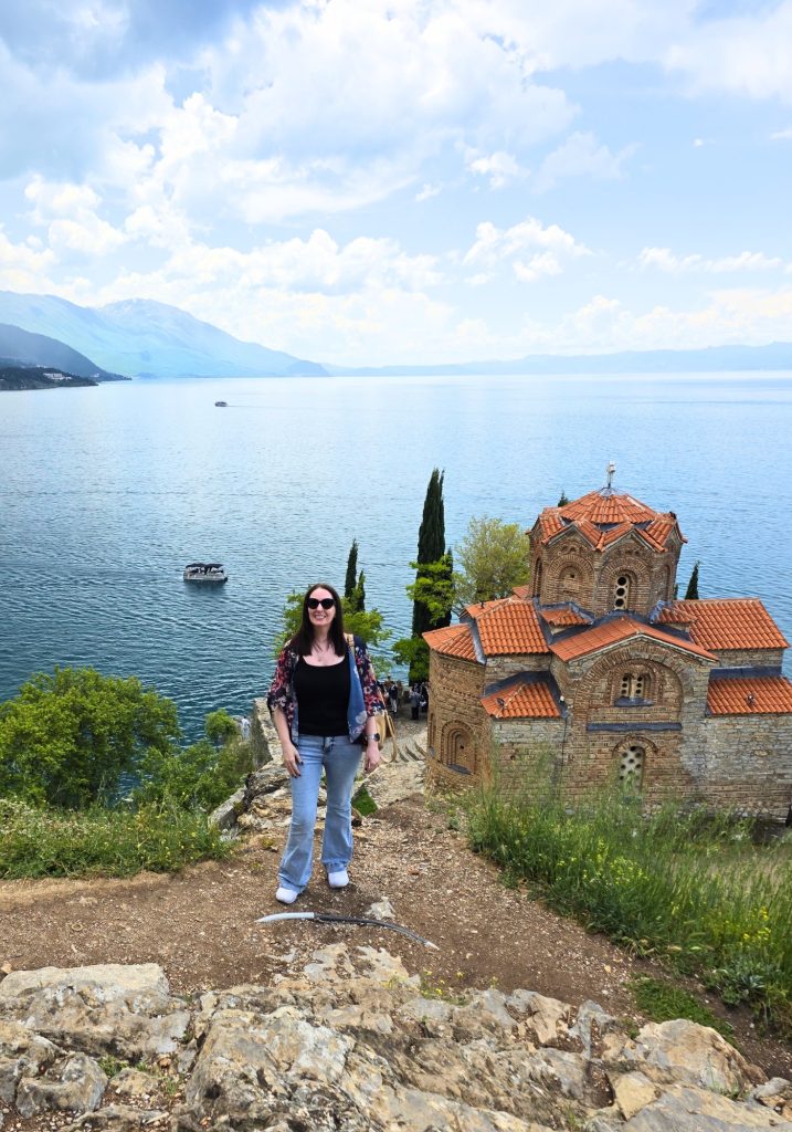 Me standing on a rocky path beside Saint John at Kaneo with Lake Ohrid and hazy mountains behind her.