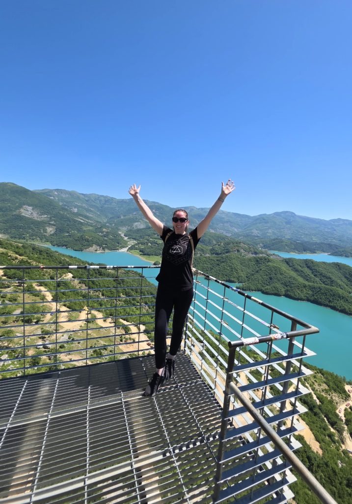 Woman stands on a metal viewing platform with both arms raised over a bright turquoise lake and green mountain valleys. Dirt roads curve below the overlook on a sunny day.