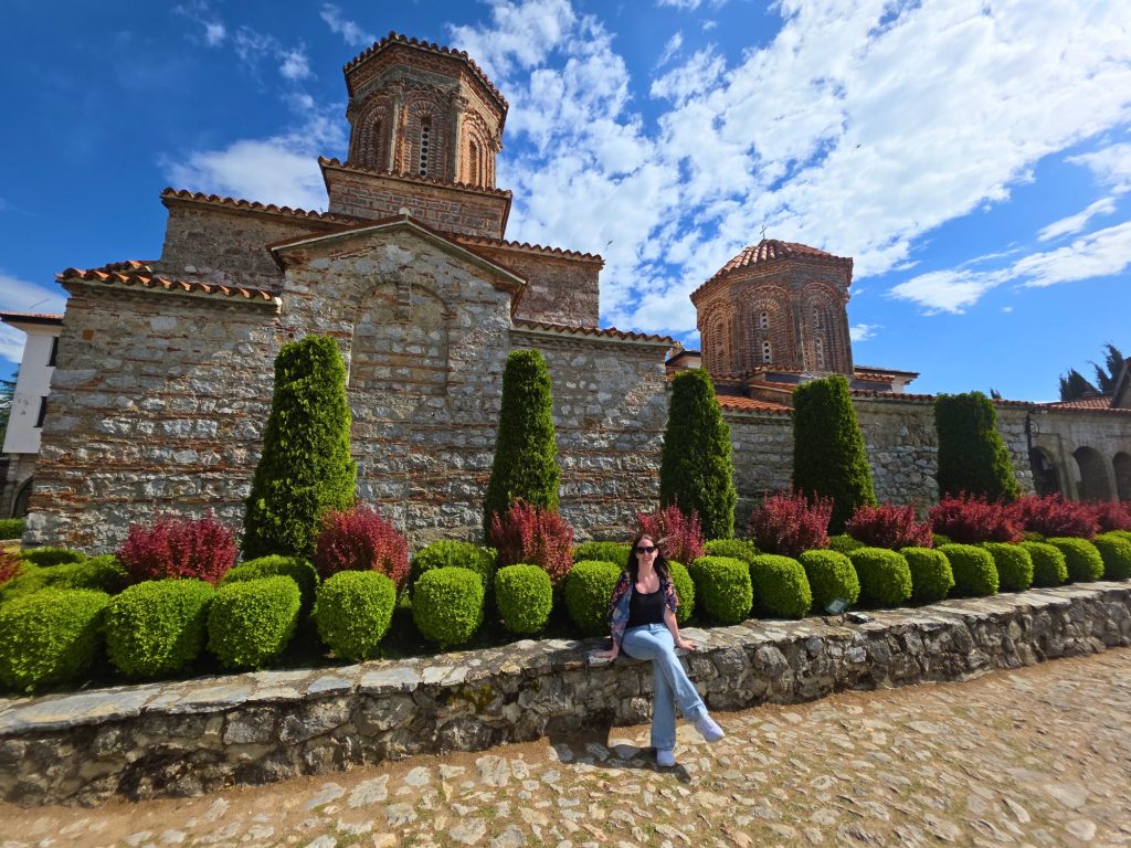 Me on a low stone wall in front of an Ohrid monastery with red domes trimmed hedges and bright blue sky above.
