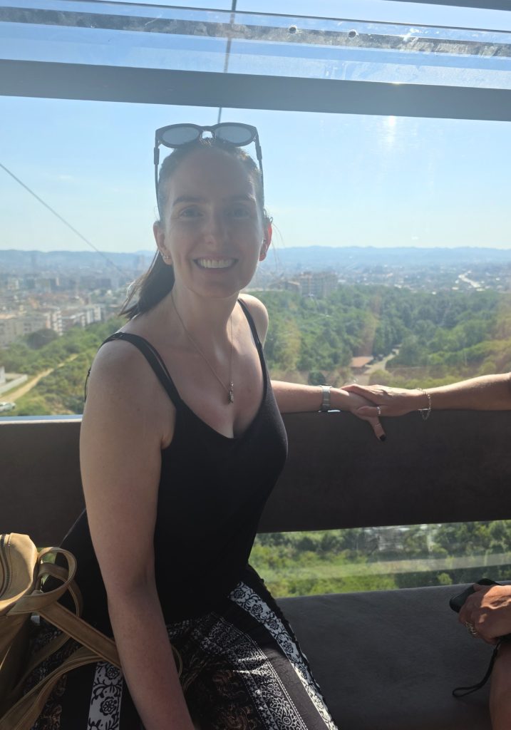 Woman smiles inside a cable car with Tirana and green hills visible through the window behind her. Bright sunlight filters through the glass as she sits beside another passenger.