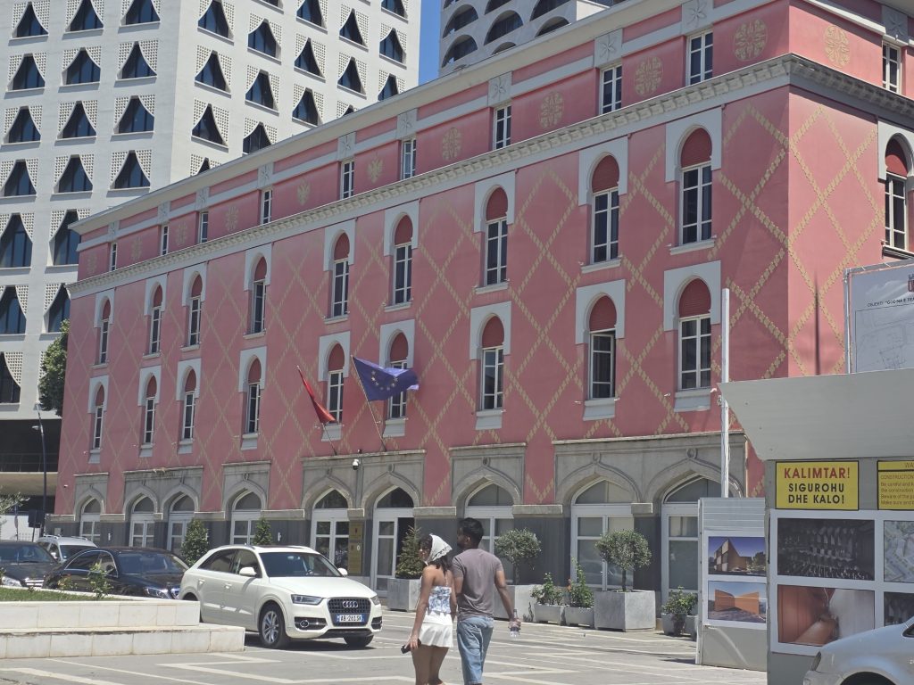 Pink historic building with arched windows and decorative gold patterns beside a modern white high rise. Albanian and European Union flags hang above the entrance while people cross the street.