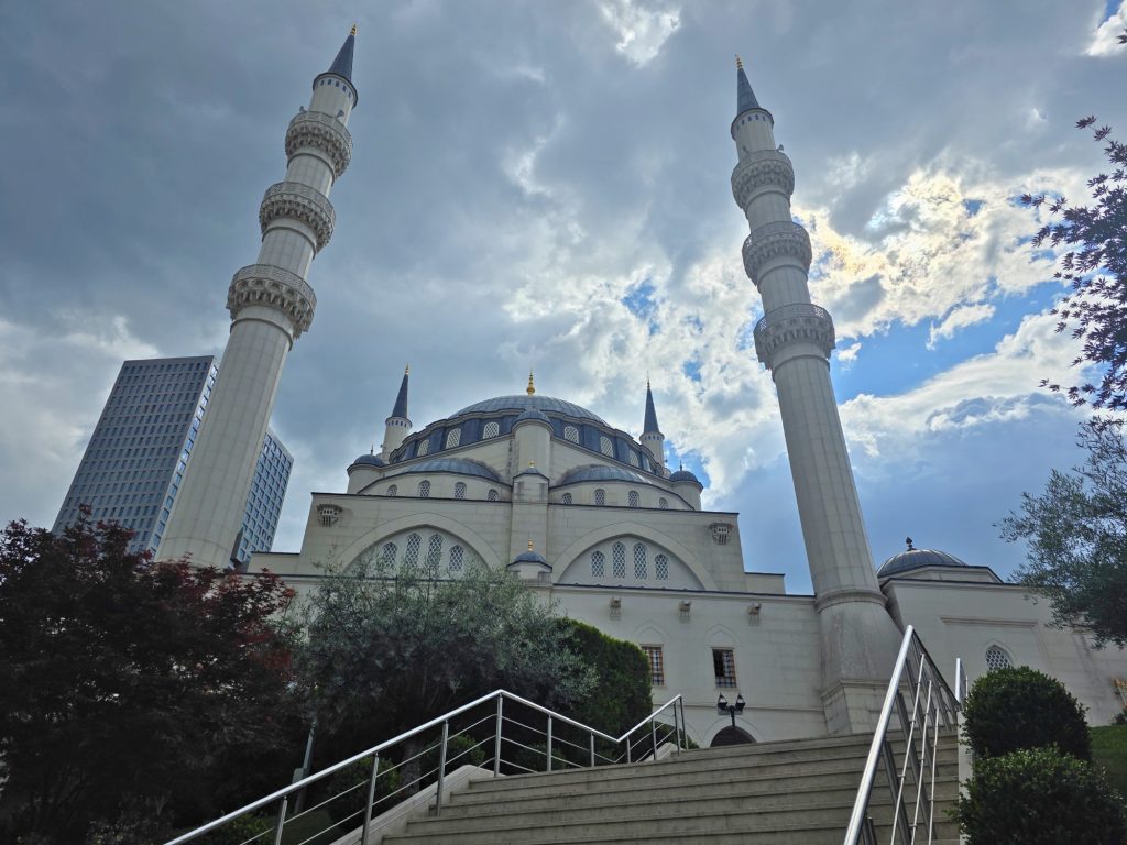 Large mosque with two tall minarets and layered gray domes viewed from the steps below. Dramatic clouds and a modern high rise rise behind the mosque.