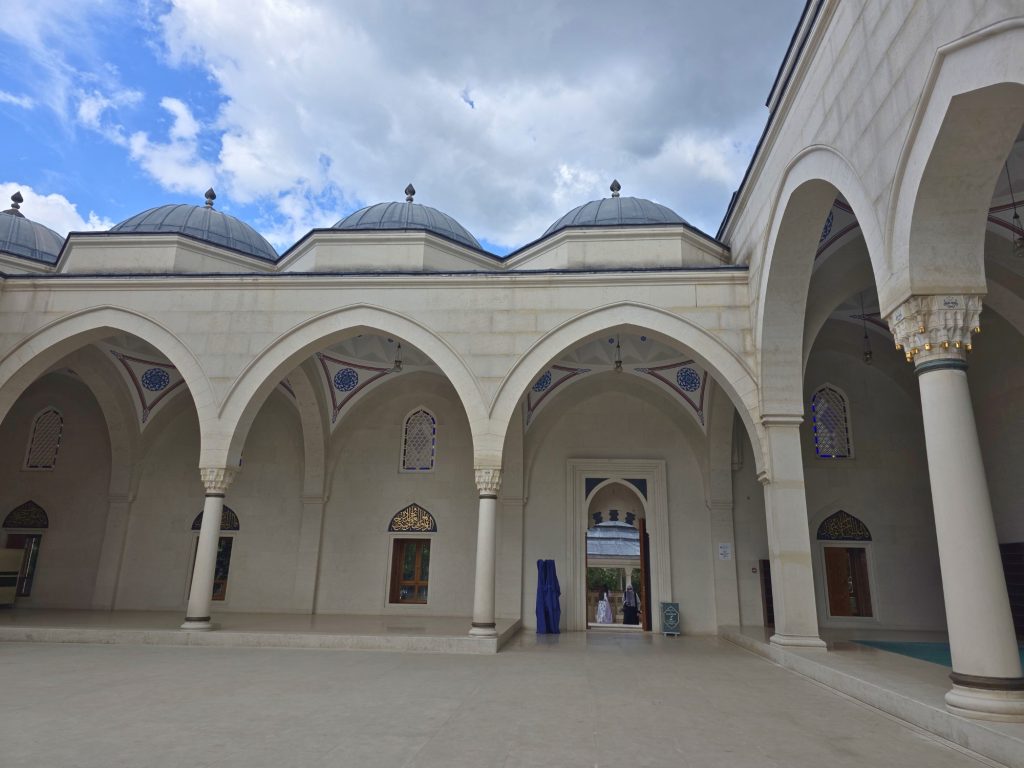 Mosque courtyard with pale stone arches. white columns. and gray domes under a cloudy sky. Blue and red decorative patterns frame the arched ceilings and windows.