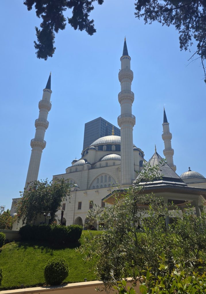 Mosque with four tall minarets and gray domes framed by trees and garden shrubs. A modern tower stands behind the mosque under a clear blue sky.