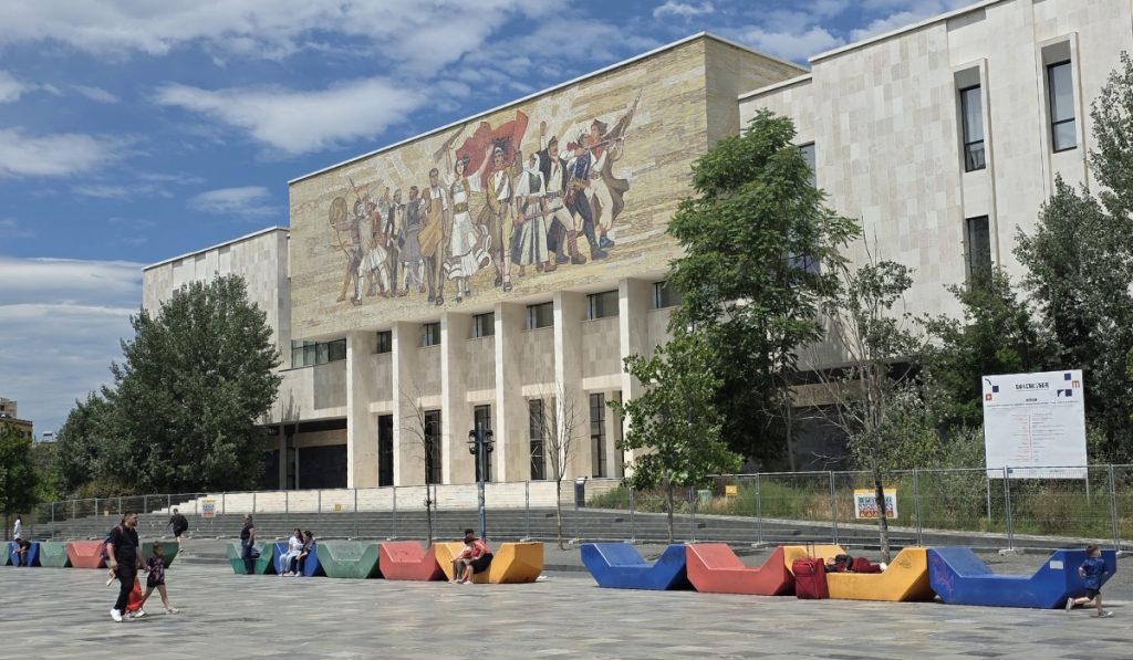 Large public building with a colorful mural of people holding flags across the upper facade. Bright geometric benches line the plaza while families and children sit and walk nearby.