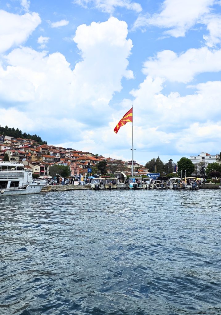 A large North Macedonian flag flies over the Ohrid waterfront with boats lined up along Lake Ohrid and hillside homes behind them.