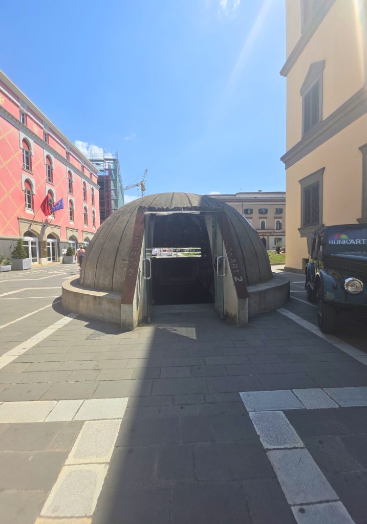 Domed concrete entrance to Bunk Art 2 between city buildings on a sunny day. The open doorway leads down into the museum and the entrance columns read "MUSEUM." and "BUNK ART 2."