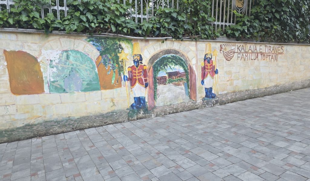 Painted mural on a stone wall showing castle arches. two toy soldiers. and an entrance path. The text reads "Kalaja e Tiranës Familja Toptani."