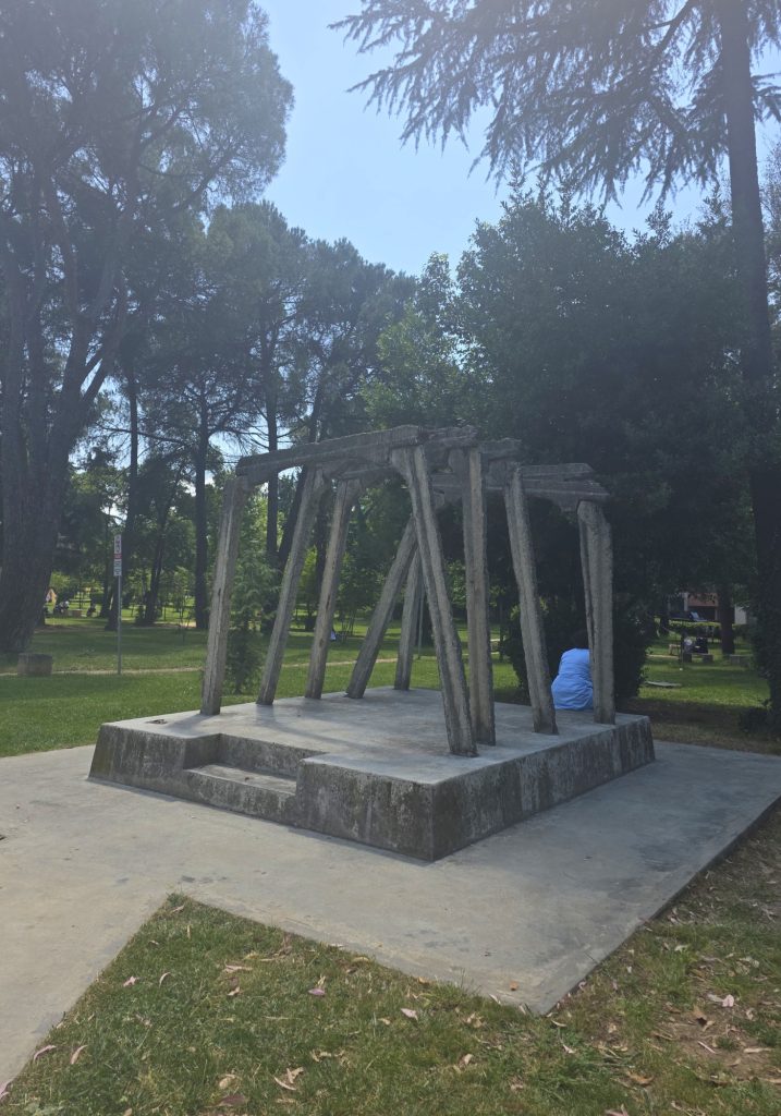 Concrete memorial sculpture shaped like unfinished beams in a shaded park. Tall trees surround the structure with benches and grass in the background.