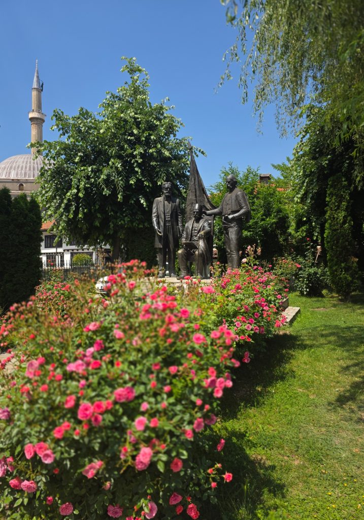 Bronze statues of three figures stand in a garden filled with bright pink flowers. A mosque dome and tall minaret rise behind the trees under a clear blue sky.