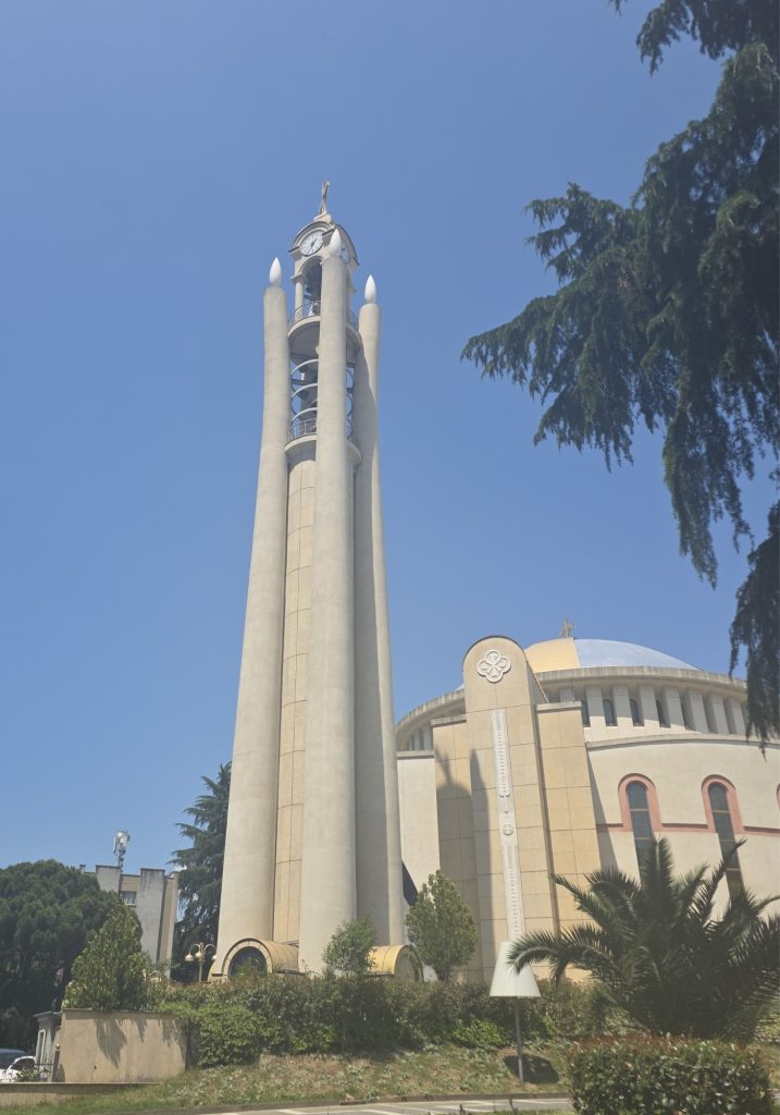 Tall church bell tower with four slender columns and a clock near the top beside a round domed church. Trees and palms surround the building under a clear blue sky.