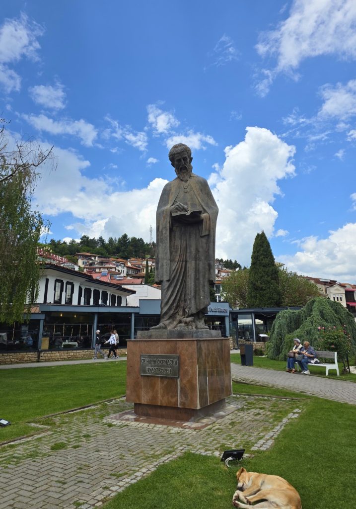 A statue of Saint Naum holding a book stands on a lawn in Ohrid with a dog resting nearby and the text "СВ. НАУМ ОХРИДСКИ ЧУДОТВОРЕЦ" on the base.