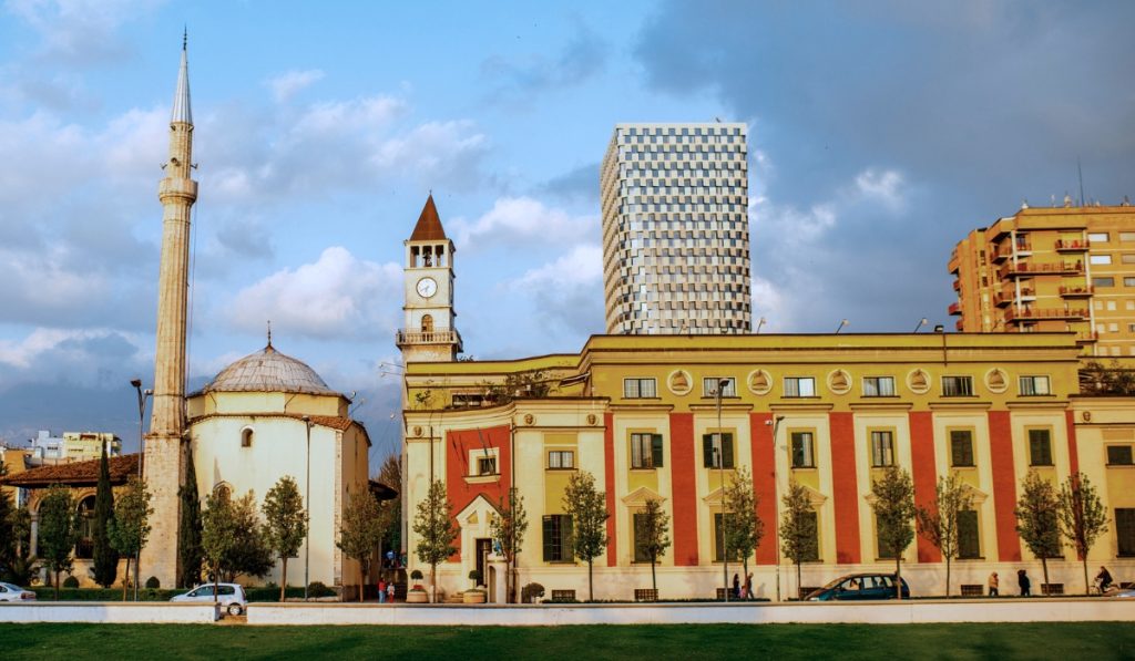 Historic mosque with a tall minaret beside a red and yellow government style building with a clock tower. A modern checkered high rise rises behind them under a dramatic cloudy sky.