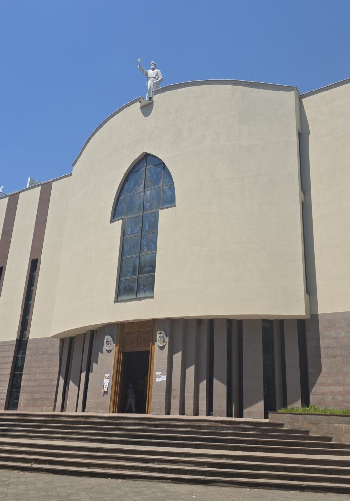 Modern church facade with a curved pale wall. tall arched stained glass window. and open wooden doors above broad front steps. A white statue stands on the roofline against the blue sky.