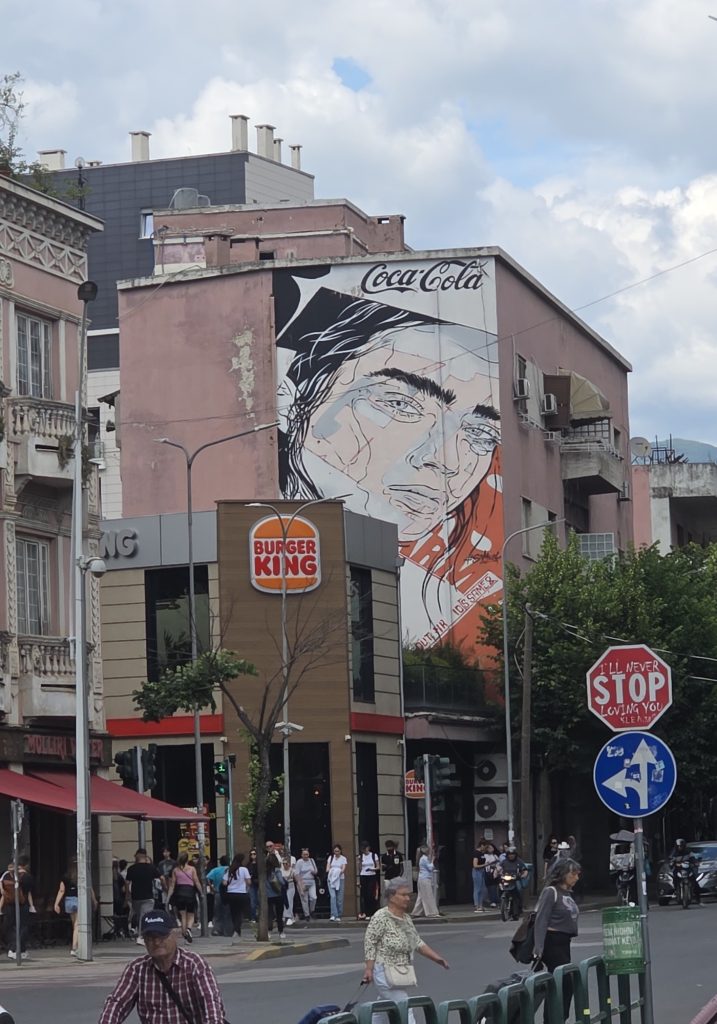 Busy Tirana street corner with a Burger King below a large Coca Cola mural of two faces on a pink building. A nearby modified stop sign reads "I’ll never stop loving you."