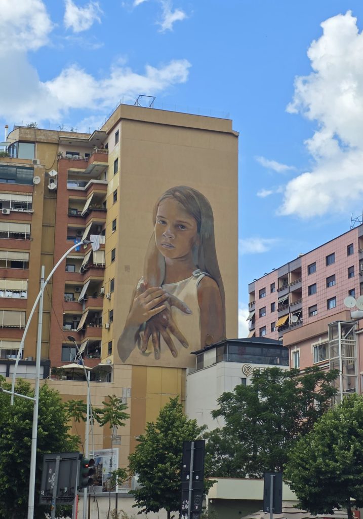 Large mural of a serious young girl painted on the side of a tall apartment building. The girl holds an oversized hand to her chest while city traffic and trees fill the street below.