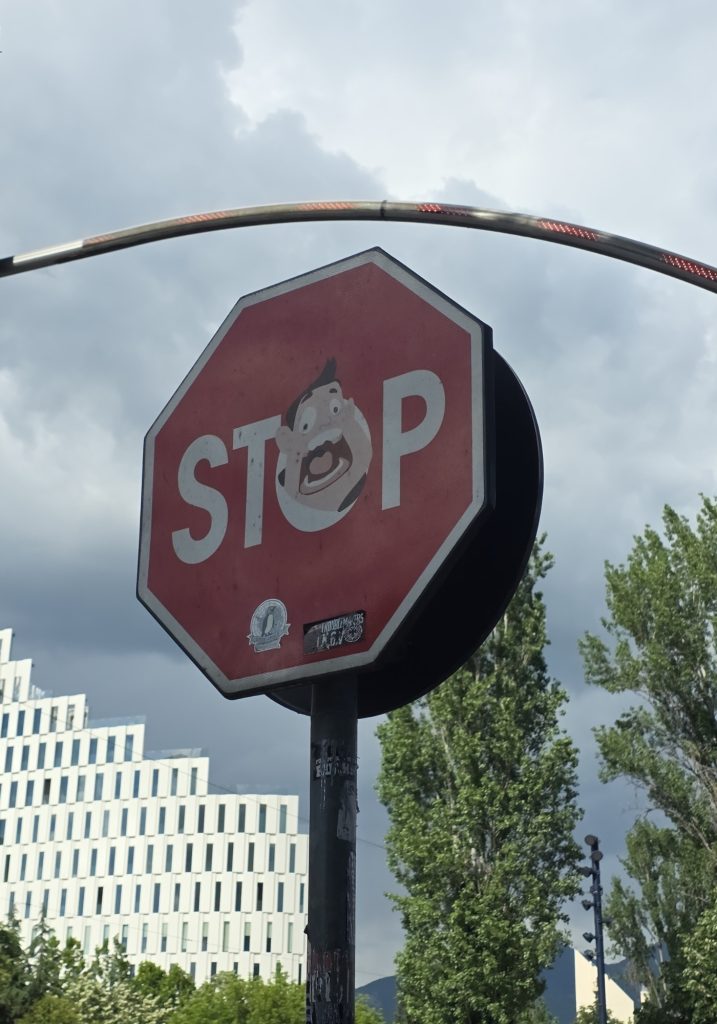 Red stop sign with a cartoon face sticker over the letter "O." Dark storm clouds. trees. and modern buildings sit behind the street sign.