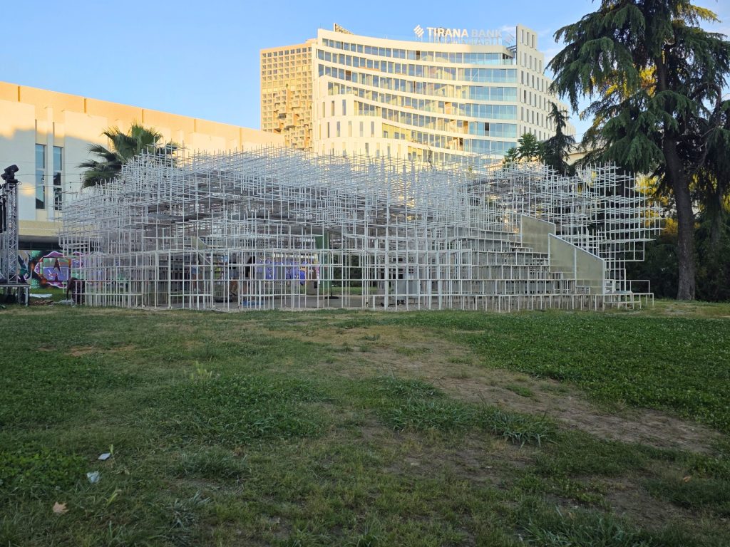 Large white grid structure on a grassy lawn with Tirana Bank buildings behind it. The open frame looks like a temporary pavilion or art installation in the evening light.