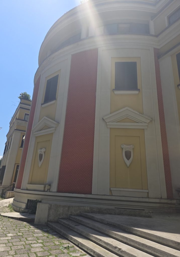 Curved historic building with red brick panels. yellow walls. dark shutters. and stone steps in bright sunlight. A strong sunbeam washes over the upper facade against a clear blue sky.
