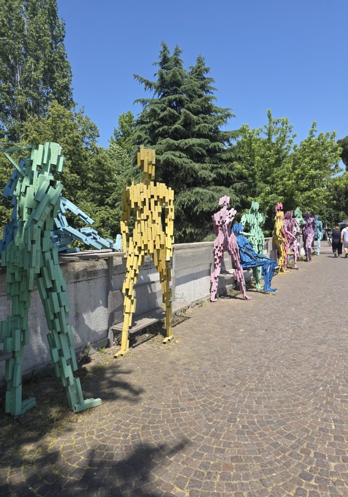 Line of colorful human shaped sculptures made from stacked metal pieces along a cobblestone path. Green trees and a clear blue sky frame the outdoor art installation.