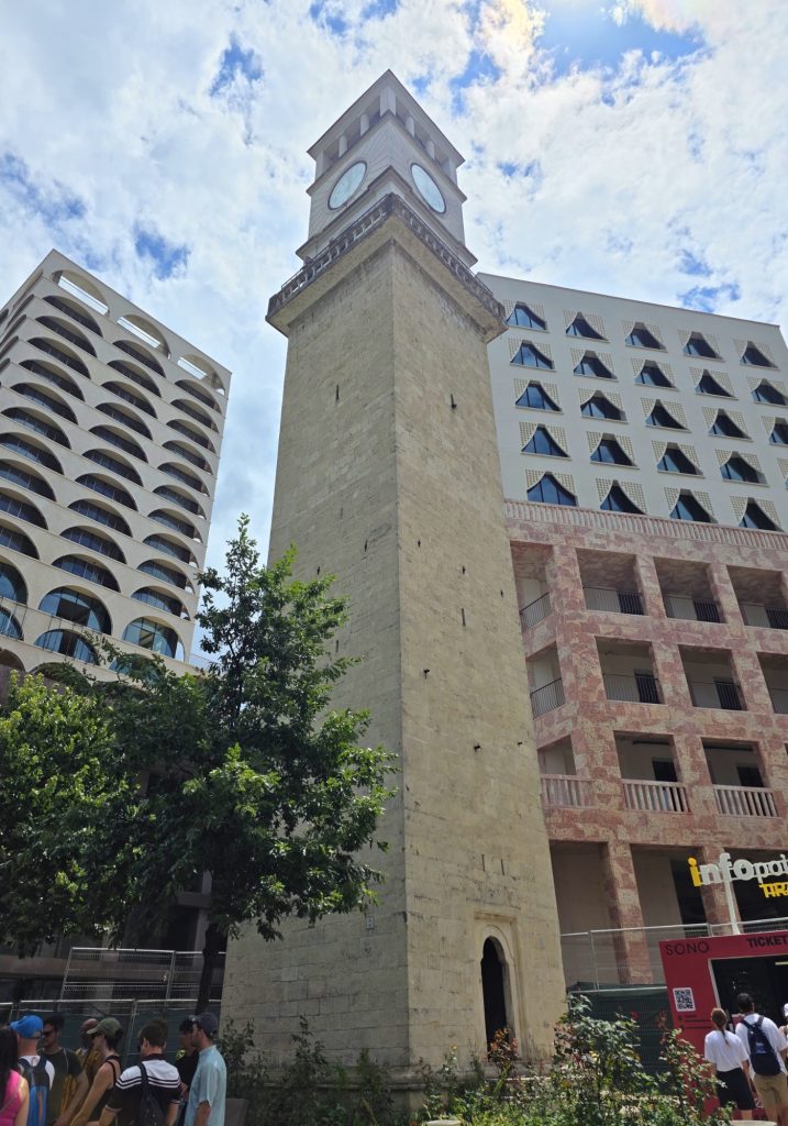 Tall stone clock tower viewed from below between modern patterned buildings. People gather near the base under a bright cloudy sky.