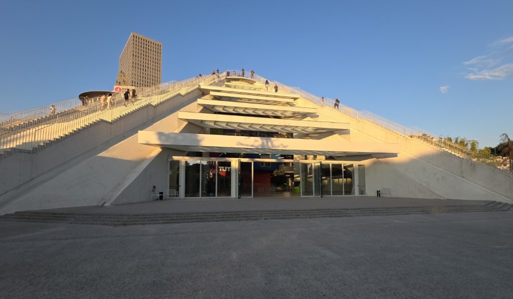Wide concrete entrance to the Pyramid of Tirana lit by golden evening sun. People walk along the staircases and upper terraces under a clear blue sky.