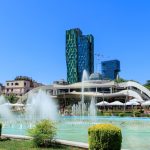 A sunny Tirana park with turquoise fountain pools spraying water in front of a white curved restaurant building. Tall green and blue modern towers rise behind it against a clear blue sky.
