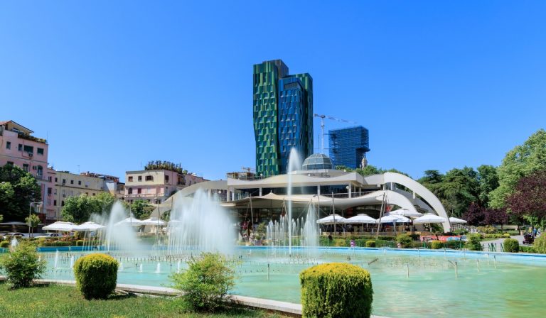 A sunny Tirana park with turquoise fountain pools spraying water in front of a white curved restaurant building. Tall green and blue modern towers rise behind it against a clear blue sky.