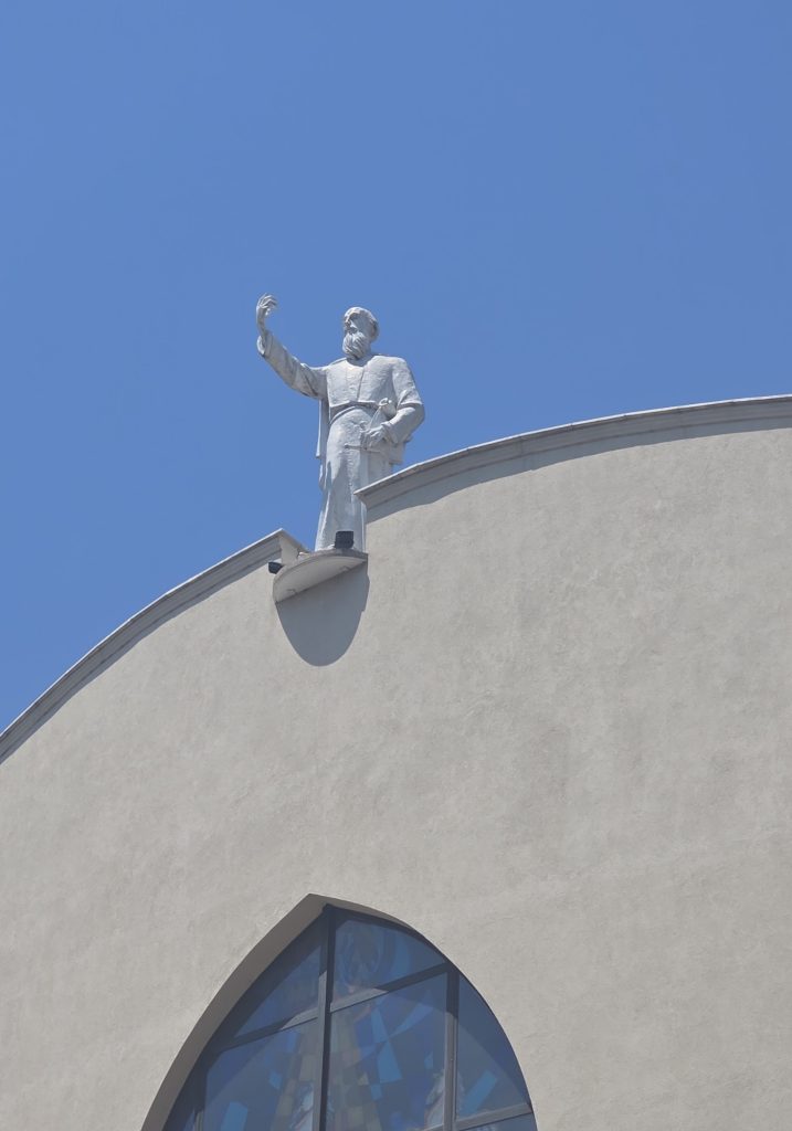 White statue of a bearded man stands above a curved church roof with one hand raised. A tall arched stained glass window sits below the statue under a clear blue sky.