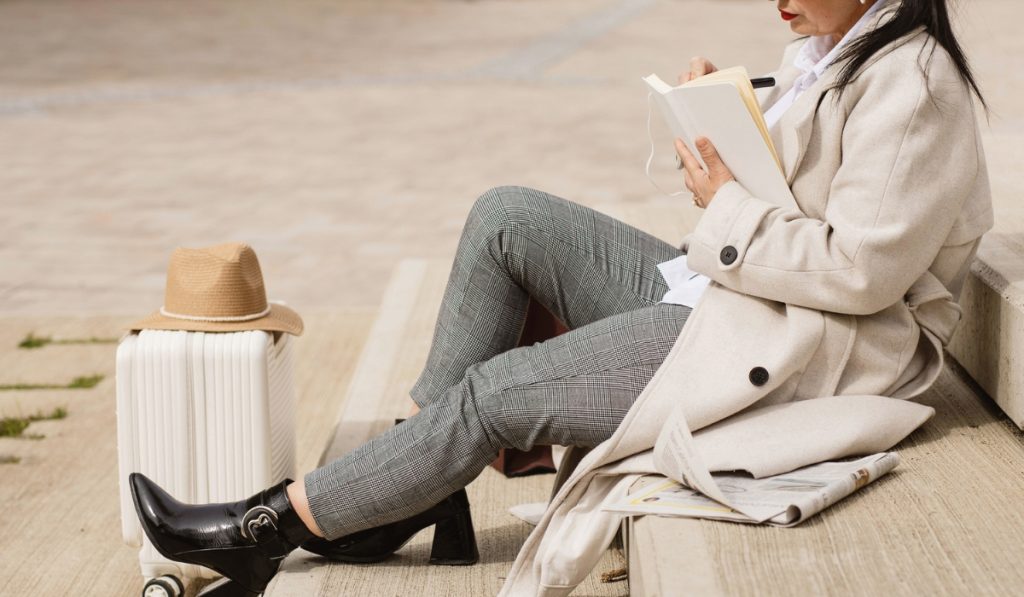 Woman sitting on outdoor steps writing in a notebook with a white suitcase and straw hat beside her. A newspaper rests on the step nearby, suggesting a quiet travel moment while waiting or journaling