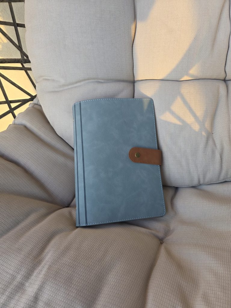 Blue travel journal with a brown snap tab resting on a cushioned wicker chair. Warm sunlight and shadows fall across the seat in a quiet outdoor setting.