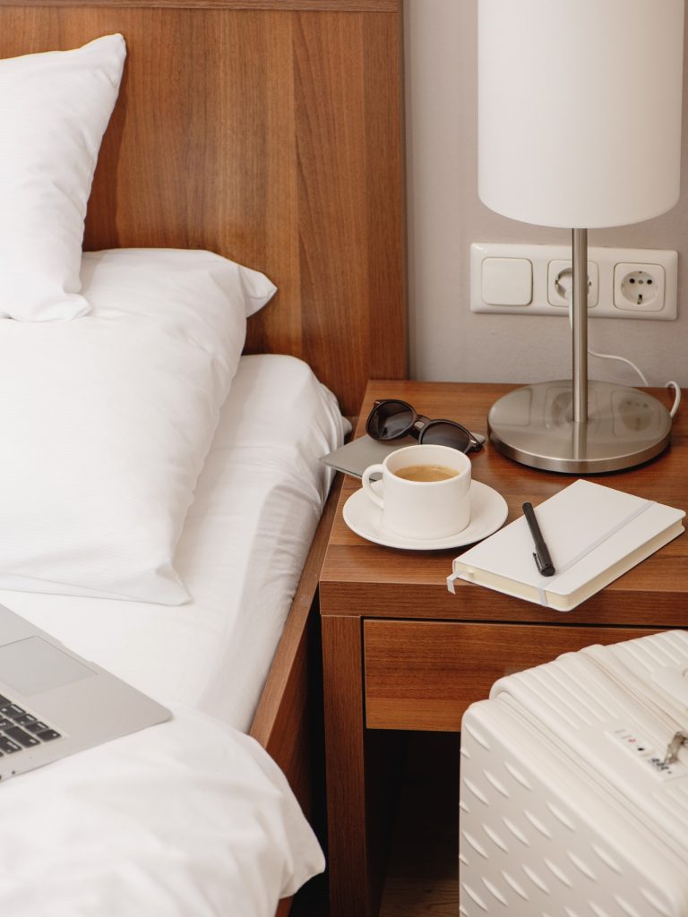 Hotel nightstand beside a white bed with coffee. sunglasses. a notebook. a pen. a lamp. and a white suitcase nearby. A laptop rests on the bed suggesting a calm work or travel planning setup.