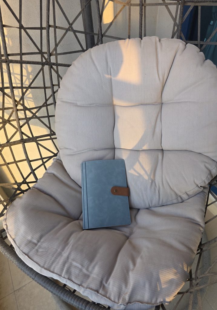 Blue travel journal with a brown snap tab resting on a cushioned wicker chair in warm sunlight. The quiet patio setup suggests a calm place for travel planning or journaling.