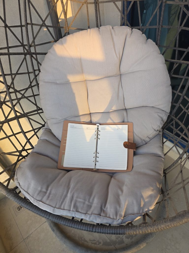 Open travel journal resting on a cushioned wicker chair in warm sunlight. The quiet patio setting suggests a peaceful place for journaling or planning during travel.