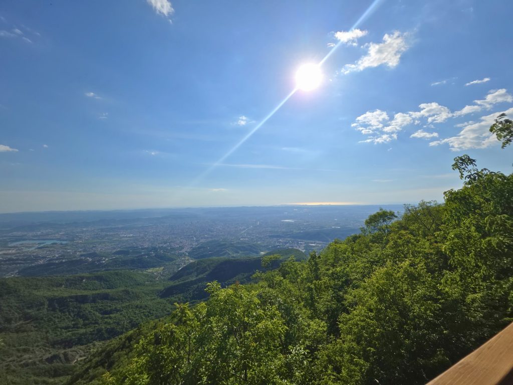 Bright sun shines over a wide view of Tirana from a mountain lookout. Green forested hills fill the foreground with the city and distant horizon stretching below.