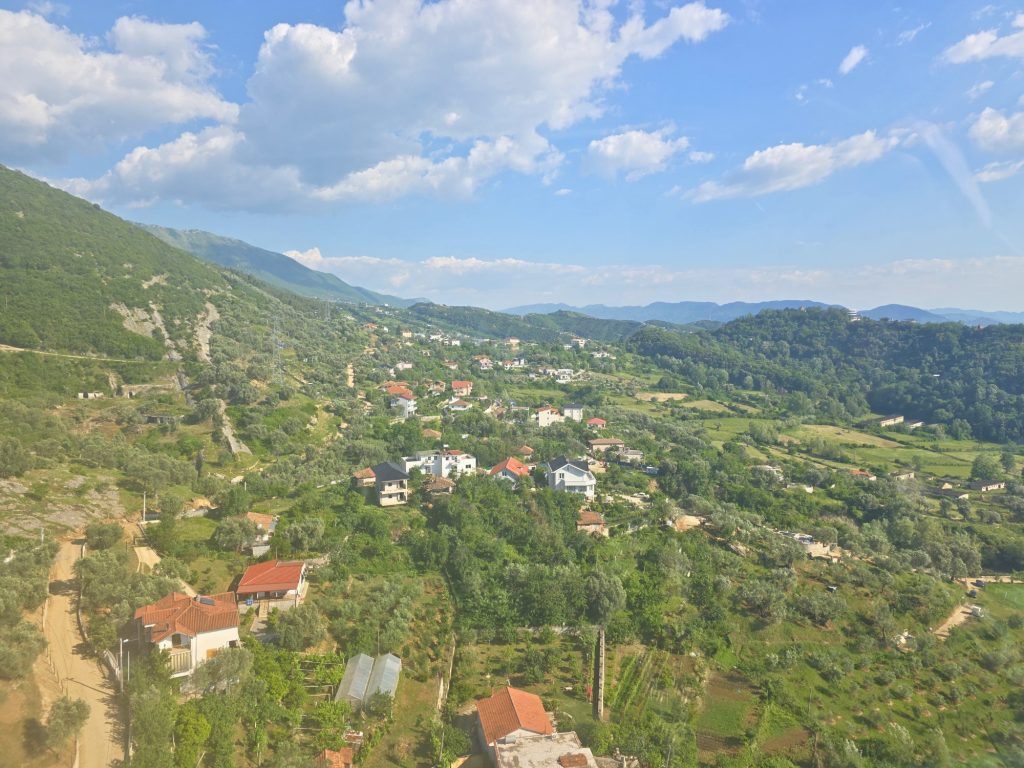 Small village with red roofed homes and garden plots spread across a green mountain valley. Forested hills and soft clouds surround the rural landscape.