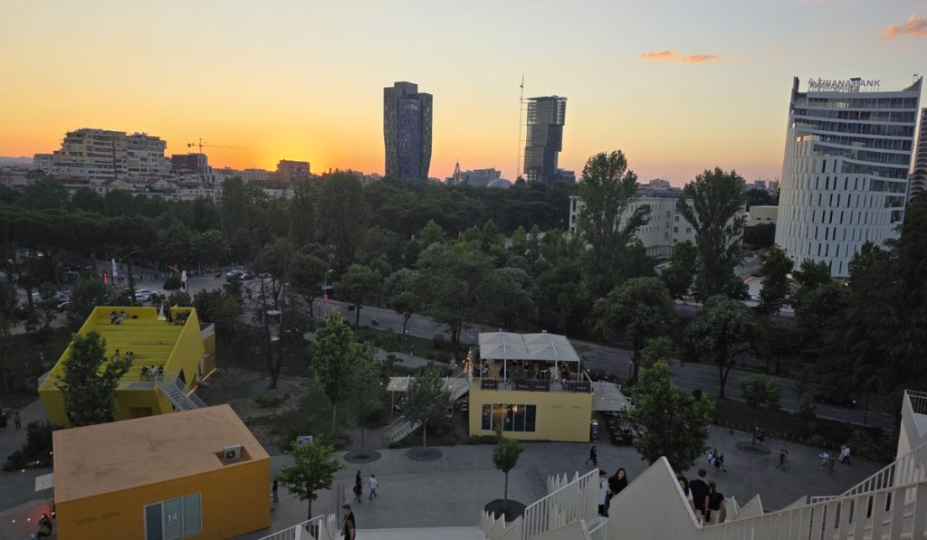 Sunset view over Tirana from an elevated terrace with yellow buildings. treetops. and modern towers on the skyline. People gather on rooftops and stairs as the sky turns orange.