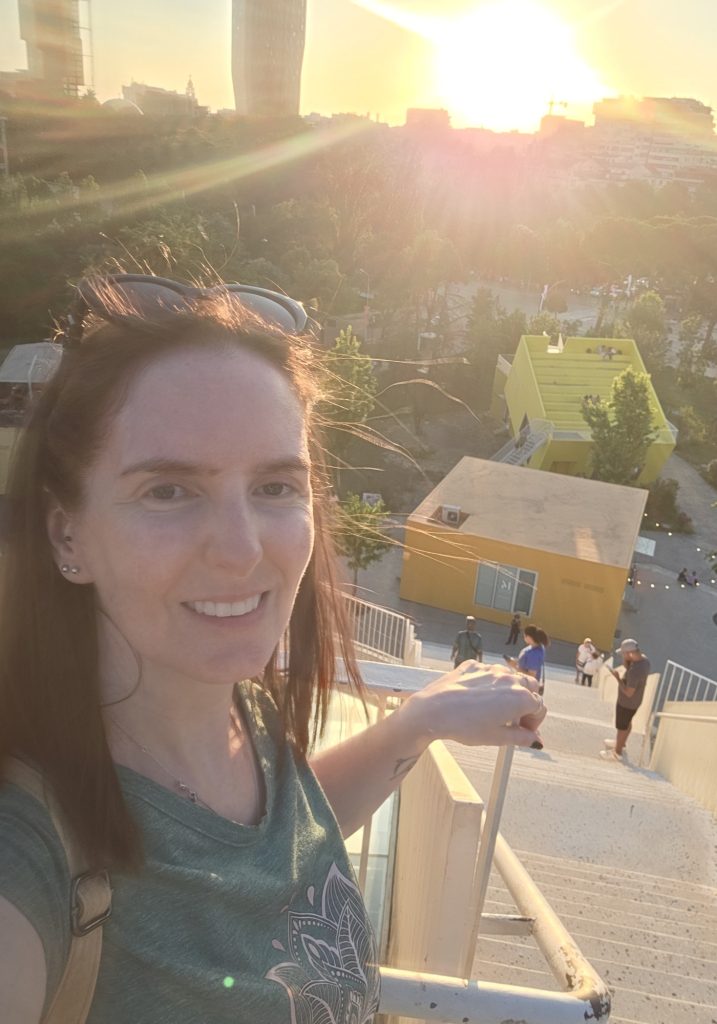 Woman takes a selfie on outdoor stairs at sunset with bright sun flare over Tirana. Yellow buildings. trees. and people walking below are visible behind her.