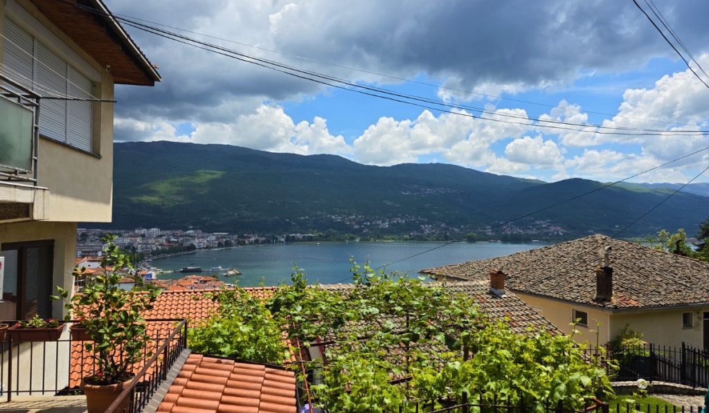 Lake Ohrid appears between red tiled rooftops and leafy grapevines with green mountains under dramatic clouds.