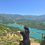Woman leans on a railing and looks out over a turquoise mountain lake from a high viewpoint. Green hills. winding dirt roads. and distant mountains fill the wide landscape.