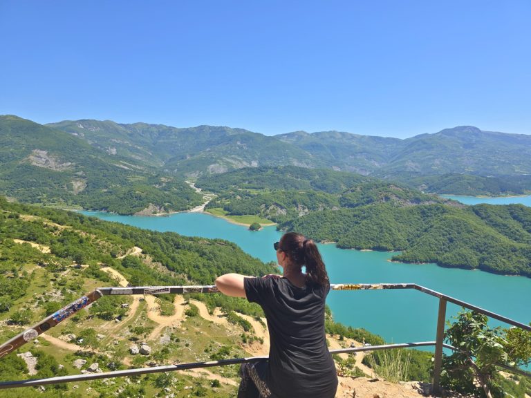 Woman leans on a railing and looks out over a turquoise mountain lake from a high viewpoint. Green hills. winding dirt roads. and distant mountains fill the wide landscape.
