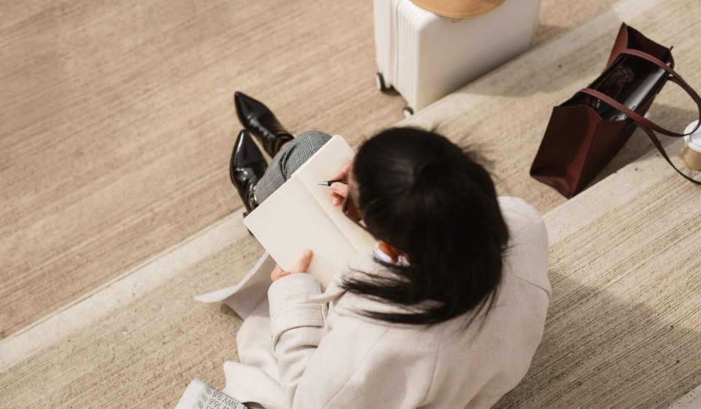 Woman sitting on outdoor steps writing in a notebook from above. A white suitcase with a straw hat and a burgundy tote sit nearby during a quiet travel moment.
