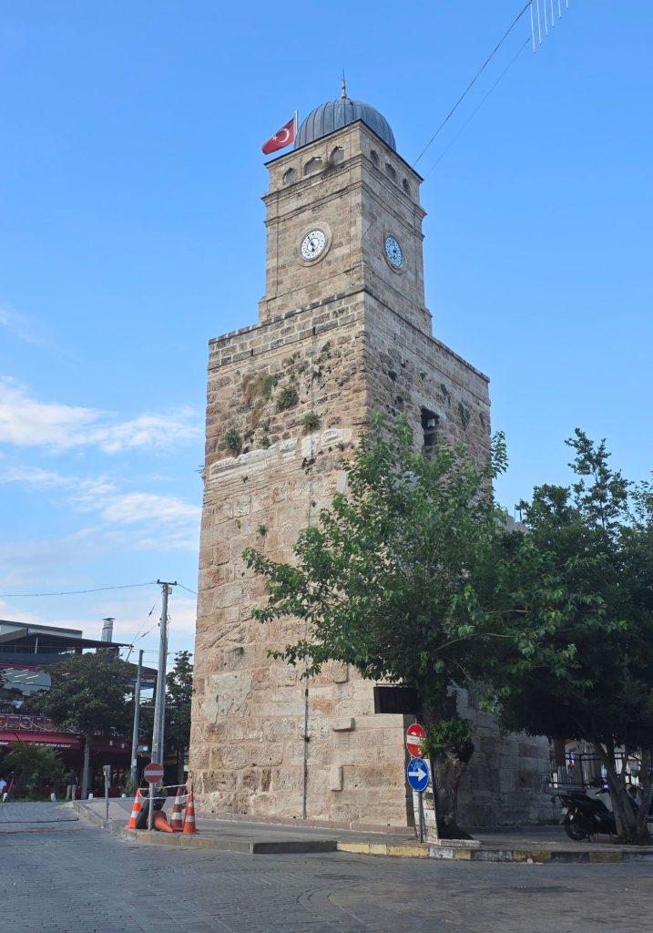 Historic stone clock tower in Antalya topped with a dark dome and a Turkish flag. Trees and quiet streets surround the landmark beneath a clear blue sky.