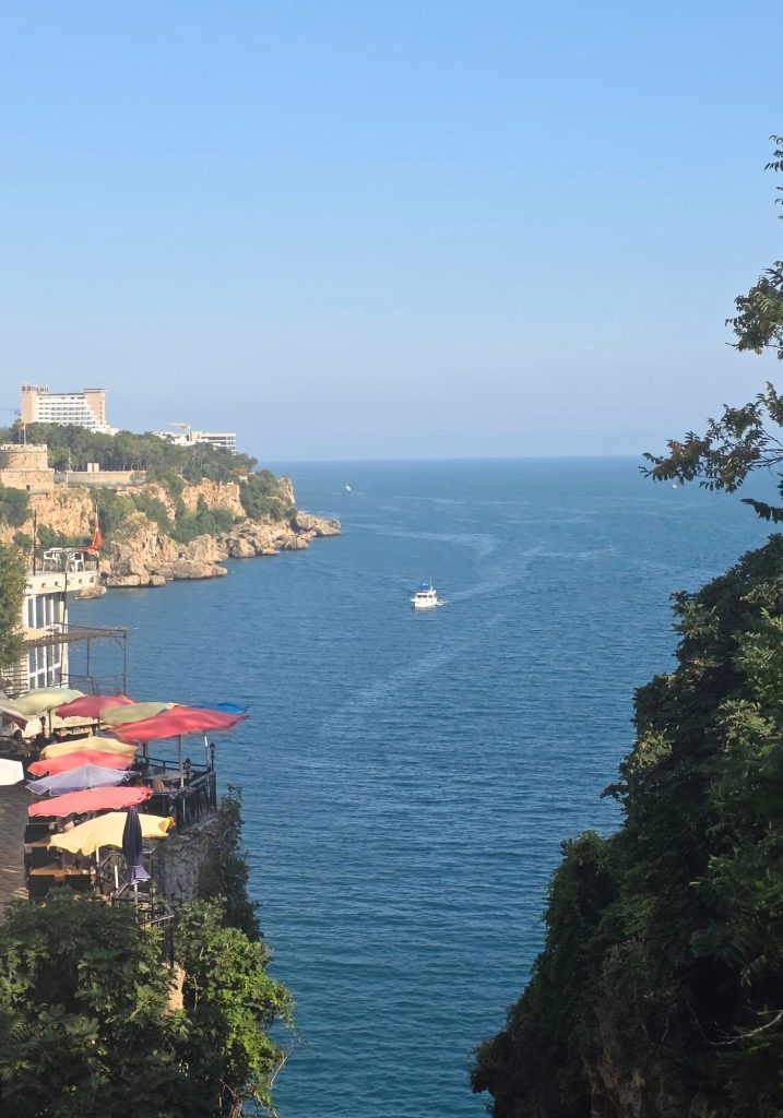 Coastal view in Antalya with a small boat on the blue Mediterranean Sea. Rocky cliffs. Seaside buildings. And a terrace shaded by colorful umbrellas frame the water.